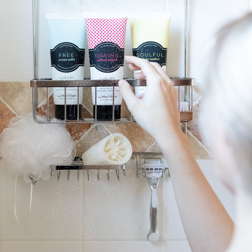 Body Wash in 3 scents (Free, Risque, Soulful) in shower on rack being reached for by blonde woman. Tile shower in background with loofah and razor