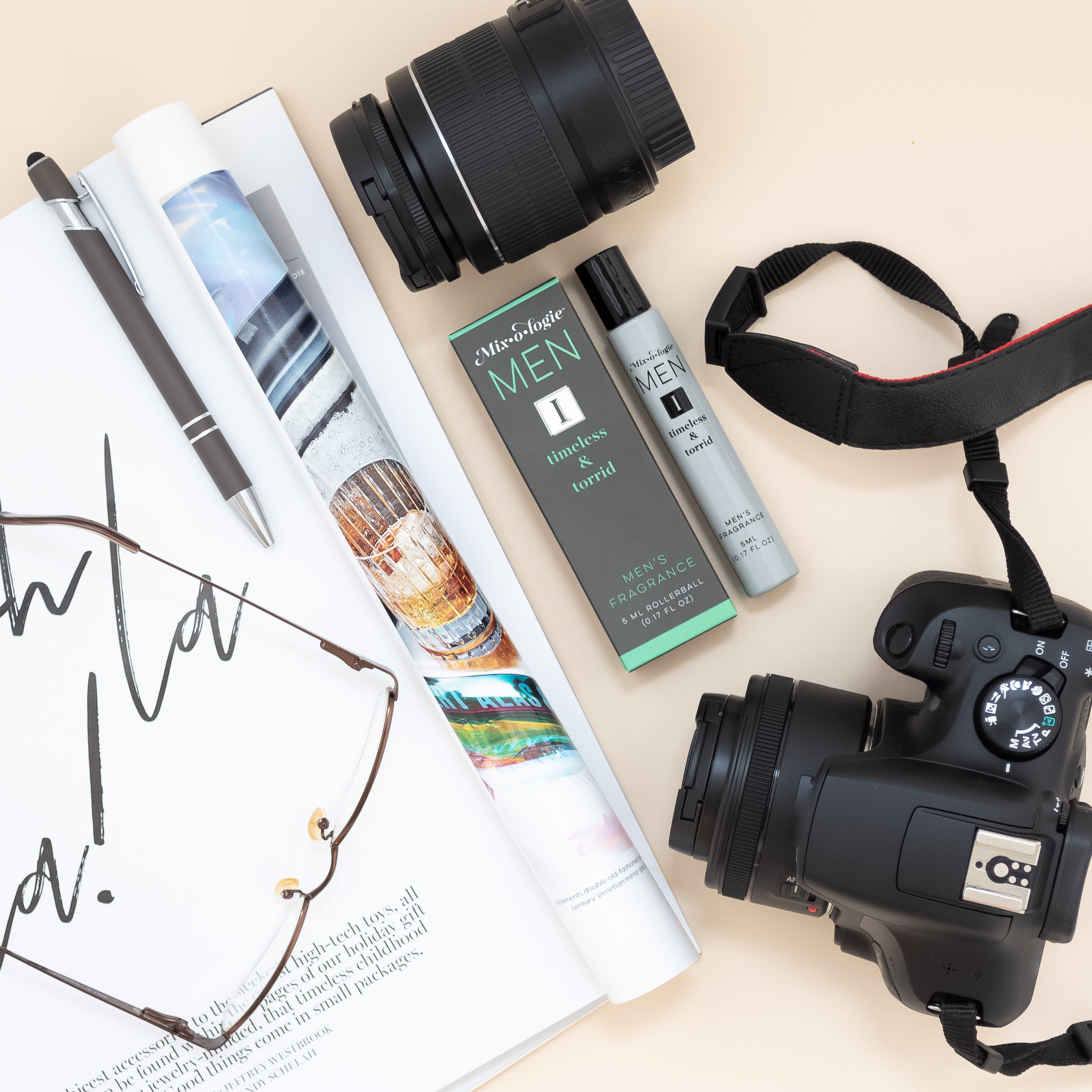 Mixologie rollerball scented with Men's I (timeless and torrid) fragrance in 5 mL glass bottle. displayed with camera, magazine, and glasses.