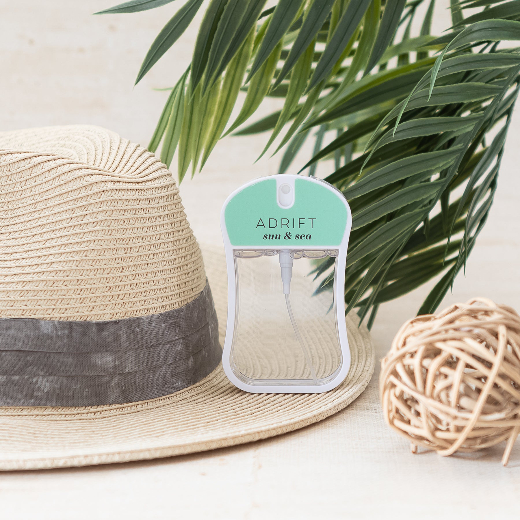 Adrift (Sun & Sea) moisturizing body mist spray bottle with light green color and clear liquid in a rounded rectangle shape. Spray bottle has 1.1 fl ox or 35 mL. spray bottle is on straw hat with palm tree in background.