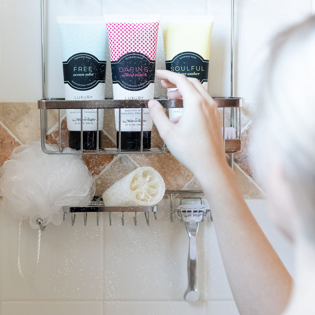 Body Wash in 3 scents (Free, Risque, Soulful) in shower on rack being reached for by blonde woman.  Tile shower in background with loofah and razor