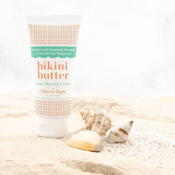 Bikini Butter Luxe Shaving cream scented with Mixologie's Tempted (coconut kiss) fragrance in 60 mL plastic tube. Displayed in sand with sea shells. 