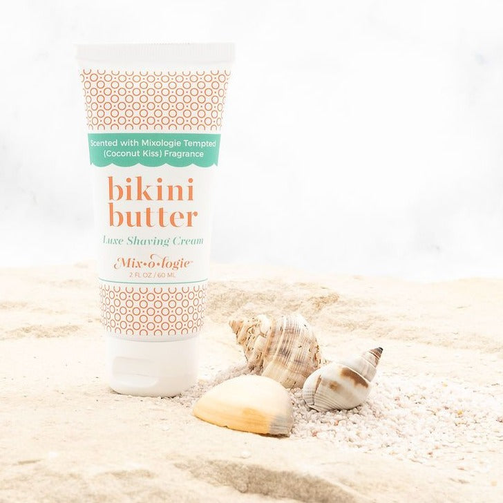 Luxe Shaving Cream Bikini Cutter scented with Mixologie Tempted (Coconut Kiss) Fragrance in a 2 fl oz or 60 mL white tube with orange circle pattern and white cap. Bikini Butter pictured in sand with sea shells.