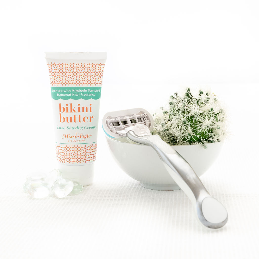 Bikini Butter Luxe Shaving cream scented with Mixologie's Tempted (coconut kiss) fragrance in 60 mL plastic tube. Displayed with cactus and shaving razor. 