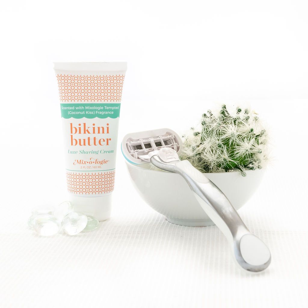 Luxe Shaving Cream Bikini Cutter scented with Mixologie Tempted (Coconut Kiss) Fragrance in a 2 fl oz or 60 mL white tube with orange circle pattern and white cap. Bikini Butter pictured with white background pebbles, a cactus, and razor.
