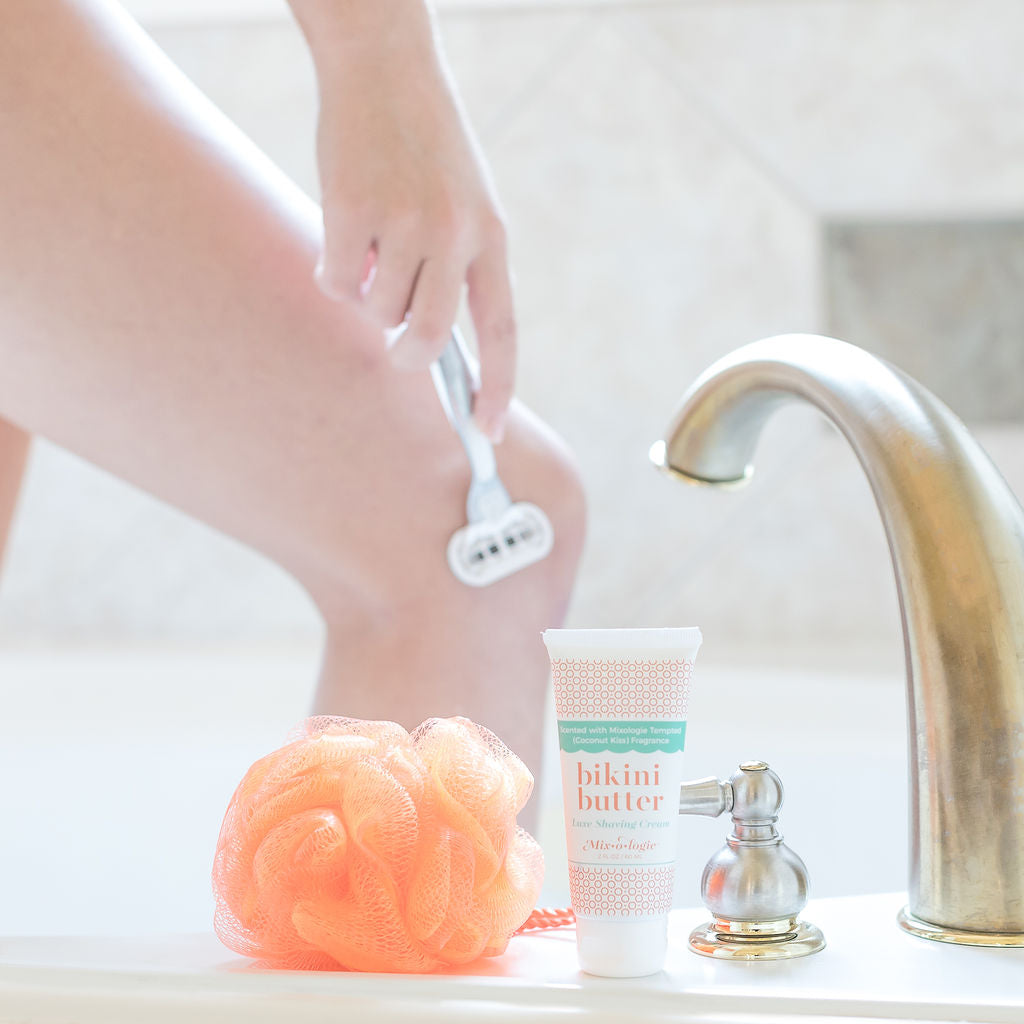 Luxe Shaving Cream Bikini Cutter scented with Mixologie Tempted (Coconut Kiss) Fragrance in a 2 fl oz or 60 mL white tube with orange circle pattern and white cap. Bikini Butter pictured on a bathtub with loofah while model shaves leg in background.