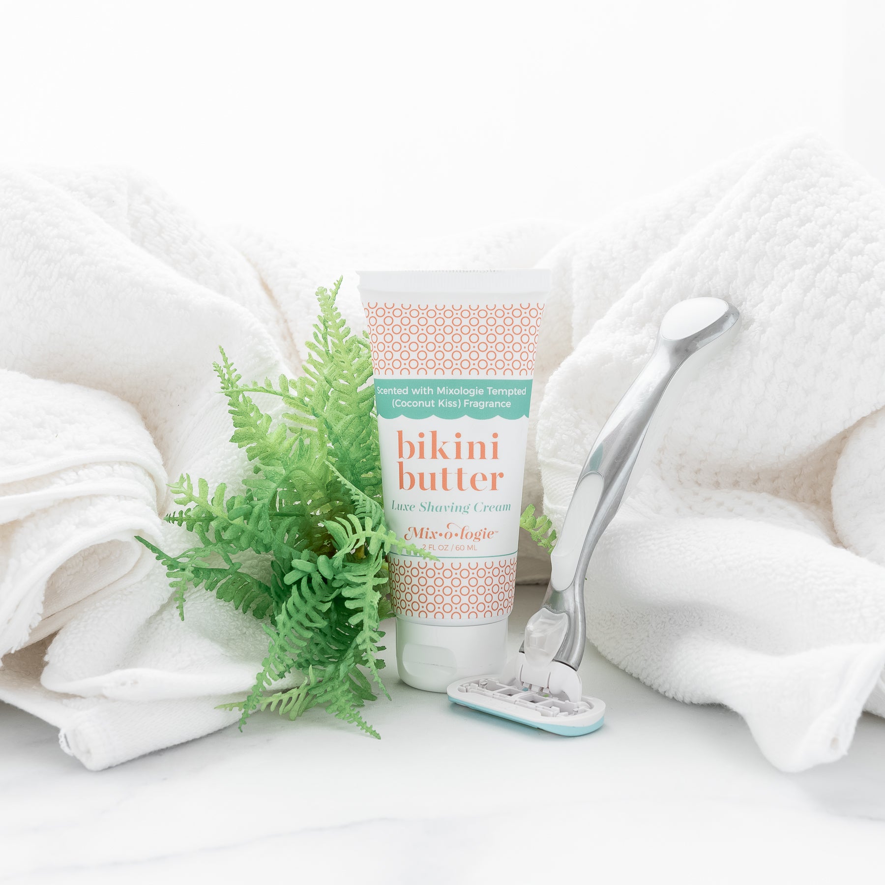 Bikini Butter Luxe Shaving cream scented with Mixologie's Tempted (coconut kiss) fragrance in 60 mL plastic tube. Displayed with shaving razor and towel. 