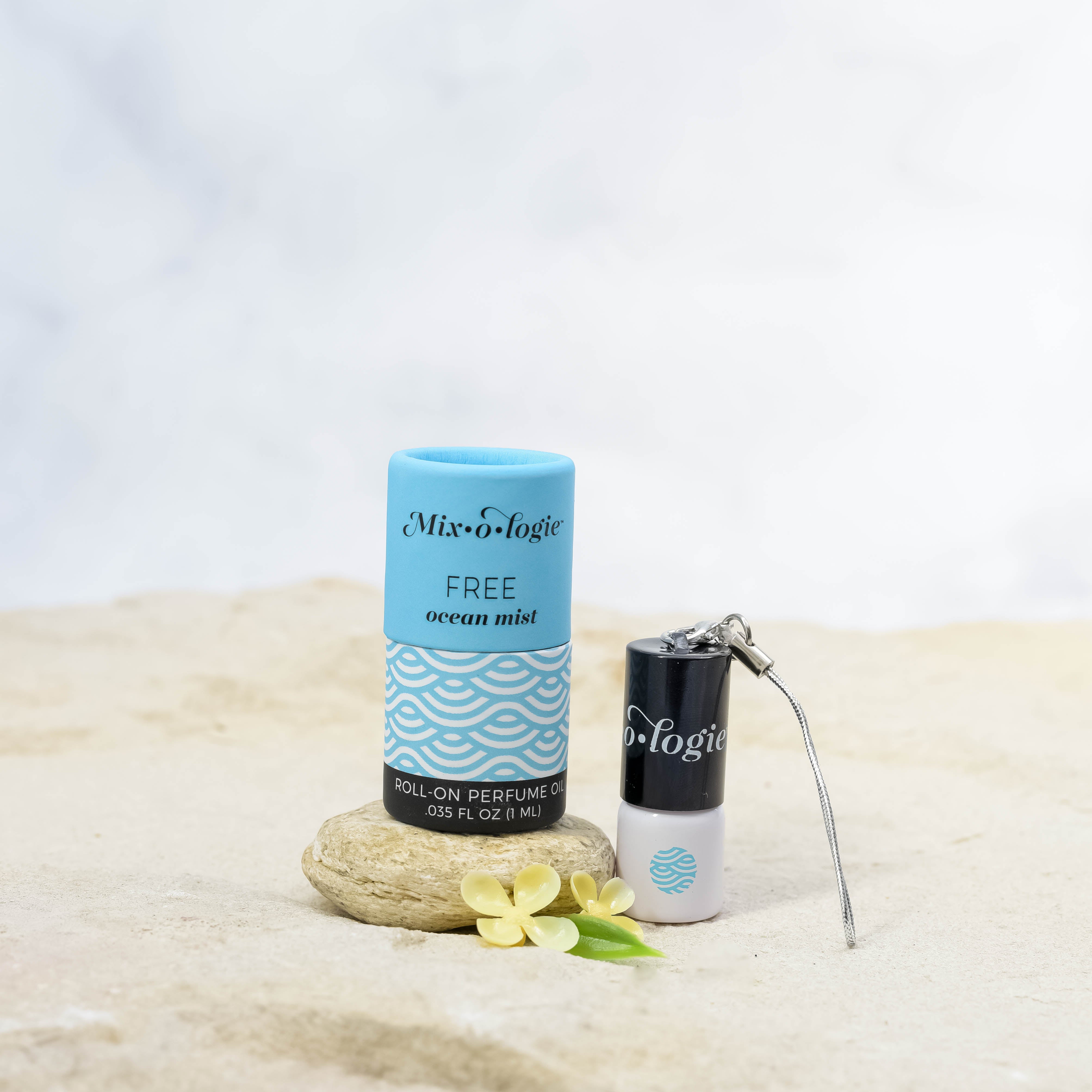Mixologie's Free (ocean mist) scented mini rollerball in 1 mL bottle with keychain lid. displayed with cylinder packaging on sand.
