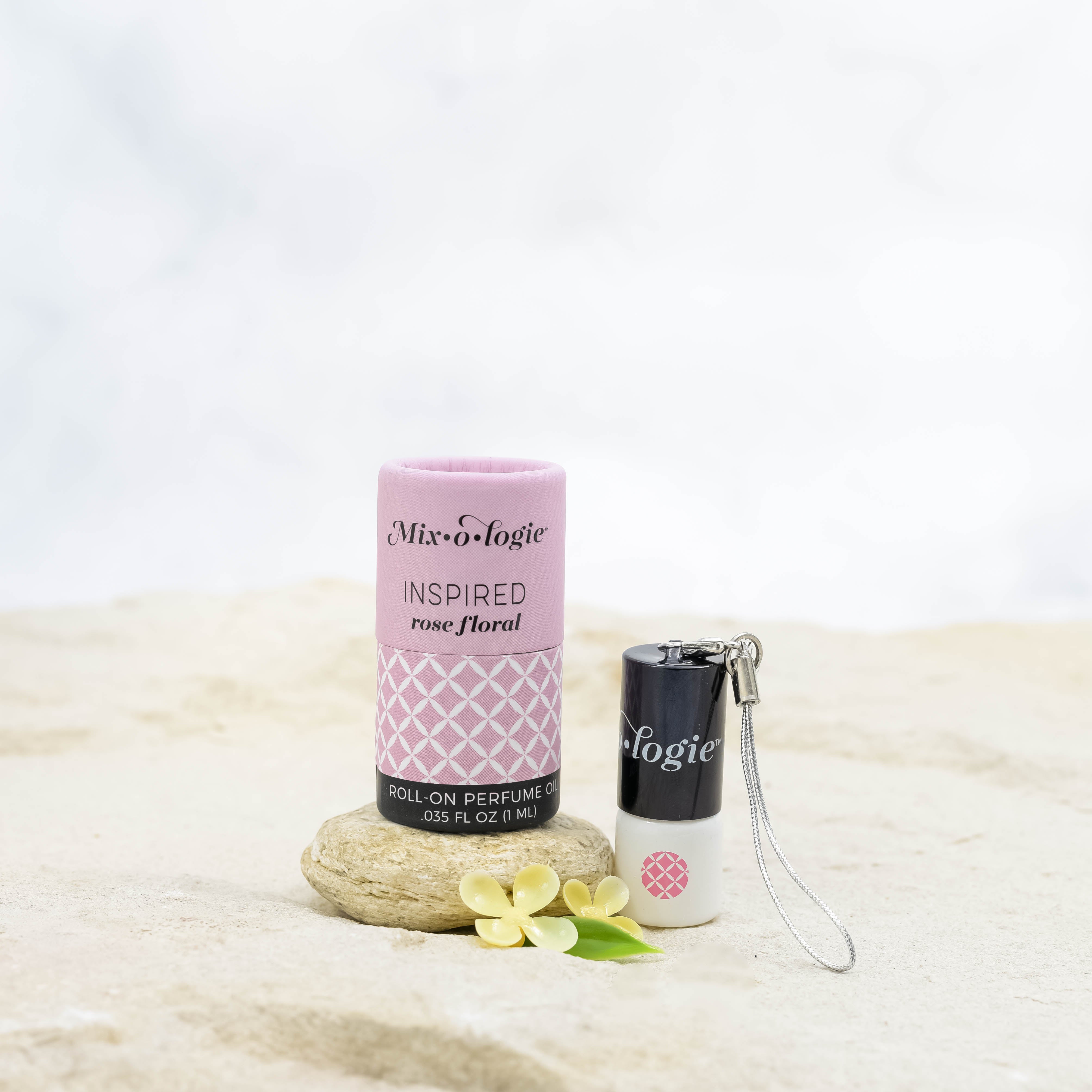 Mixologie's Inspired (rose floral) scented mini rollerball in 1 mL bottle with keychain lid. displayed with cylinder packaging on sand.