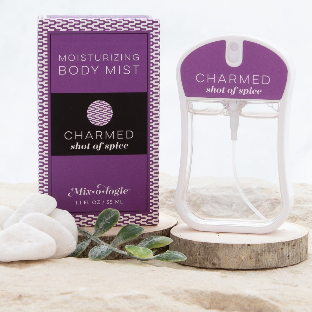 Charmed (Shot of Spice) Moisturizing Body Mist in dark purple color box and rounded rectangle spray bottle with dark purple color label and clear liquid. Spray bottle has 1.1 fl oz or 35 mL. Spray bottle and box are pictured on wood in sand with pebbles and greenery.