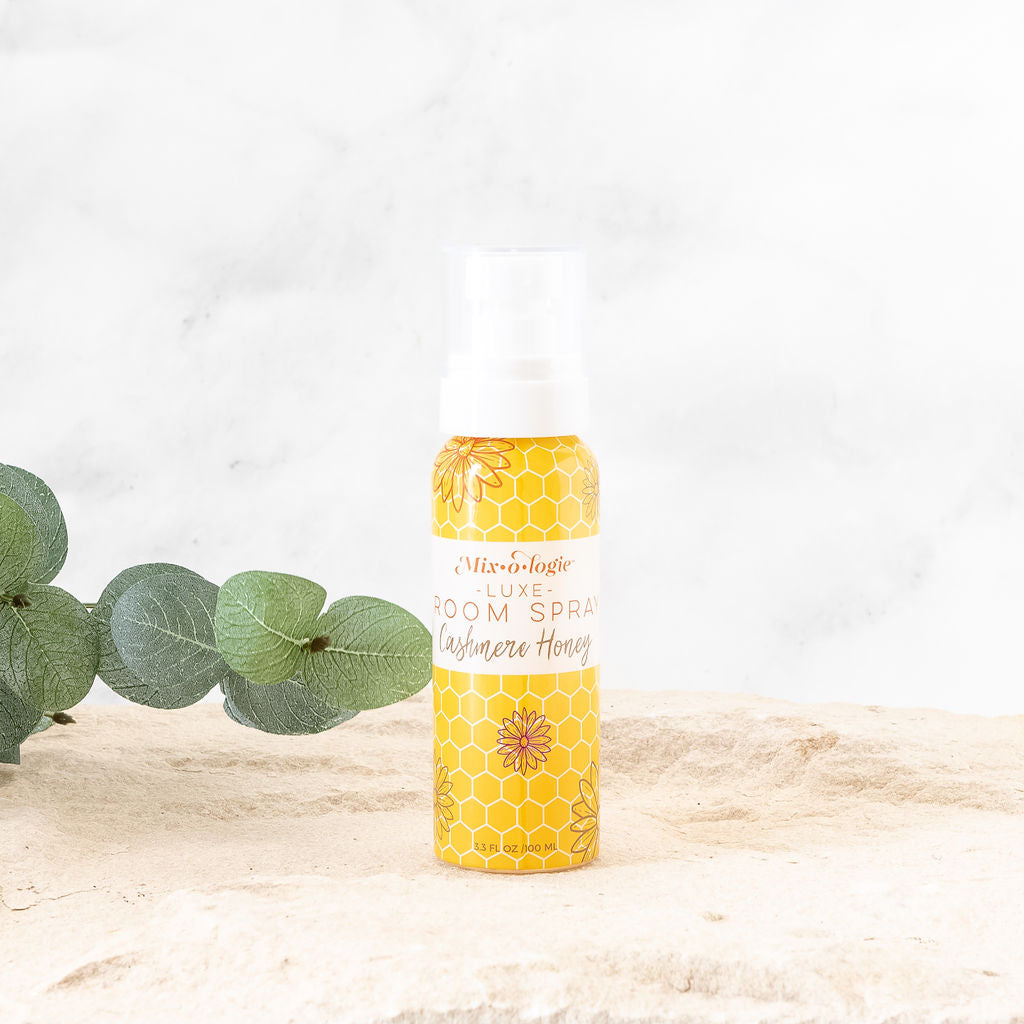Luxe Room Spray in Mixologie’s scent Cashmere Honey in a 3.1 fl oz or 100 mL yellow honeycomb and flower cylinder spray bottle with white label and white spray top. Pictured in sand with greenery. 