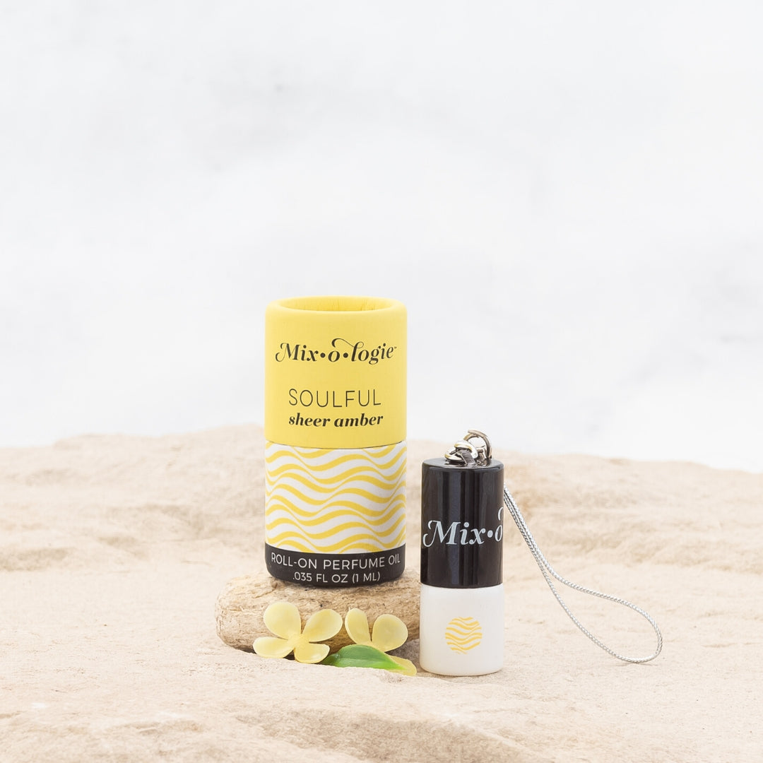 Mixologie's Soulful (sheer amber) scented mini rollerball in 1 mL bottle with keychain lid. displayed with cylinder packaging on sand.