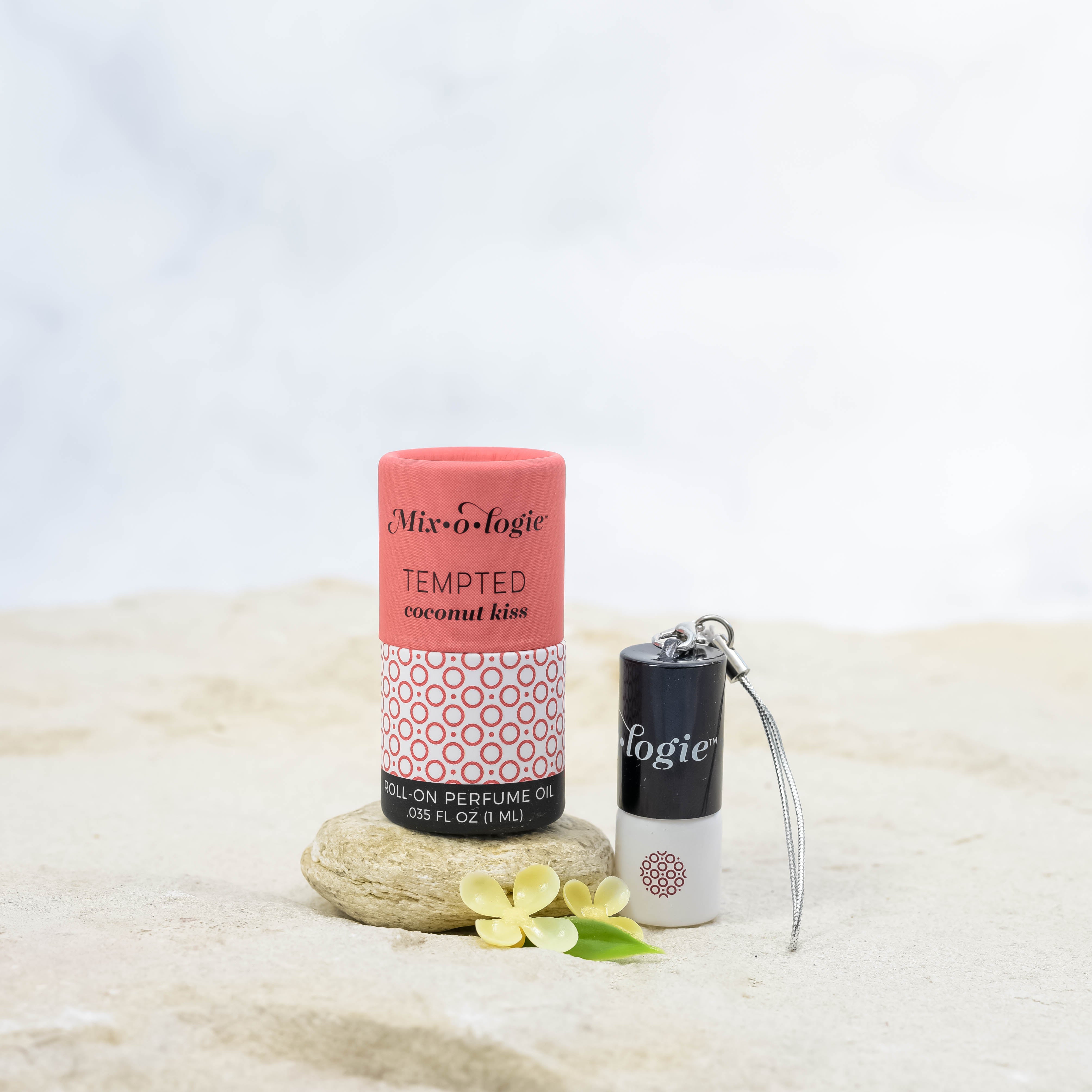 Mixologie's Tempted (coconut kiss) scented mini rollerball in 1 mL bottle with keychain lid. Displayed with cylinder packaging on sand.