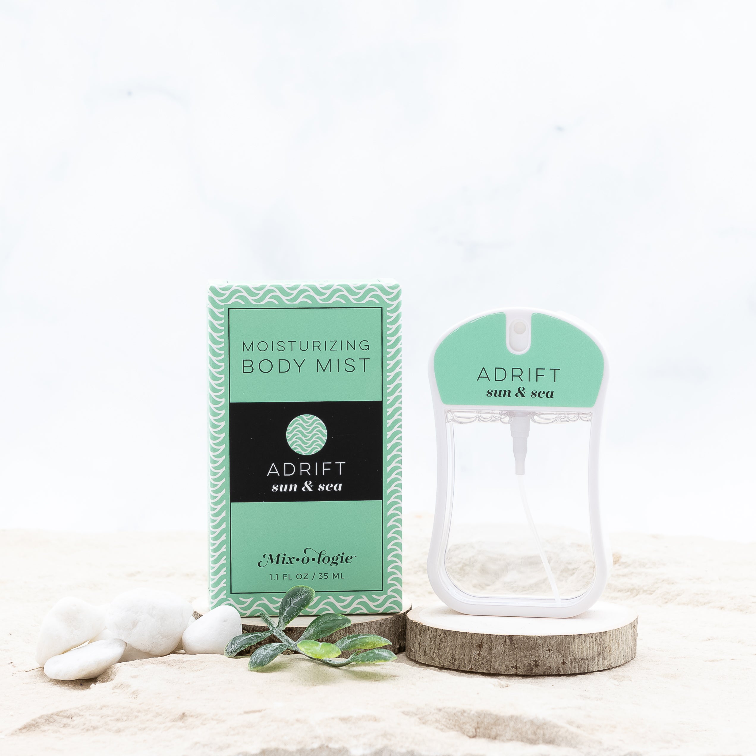 Adrift (Sun & Sea) moisturizing body mist in light green color box and spray bottle with light green color label and clear liquid. Spray bottle has 1.1 fl oz or 35 mL. Box and spray bottle are on wood in sand with greenery and pebbles.